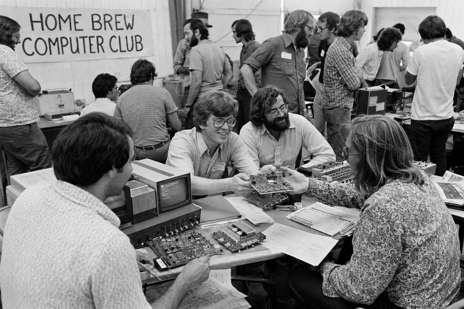 The Homebrew Computer Club, 1975 — a crowded room of people passing circuit boards across folding tables